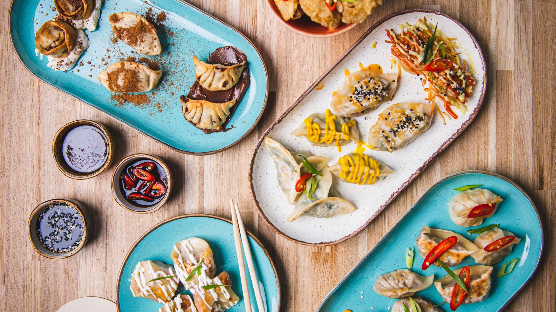 Several plates of dumplings with different toppings and sauces arranged on a wooden table alongside dipping sauces and chopsticks.