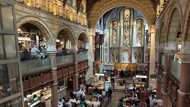 A view of the interior of Mercato Mayfair, with tables full of diners and street food vendors on the ground floor, architectural arches and stained glass windows.