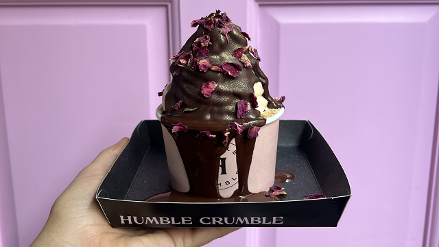 A hand holding a round pot of crumble in a square tray, drizzled in chocolate sauce and scattered with edible rose petals