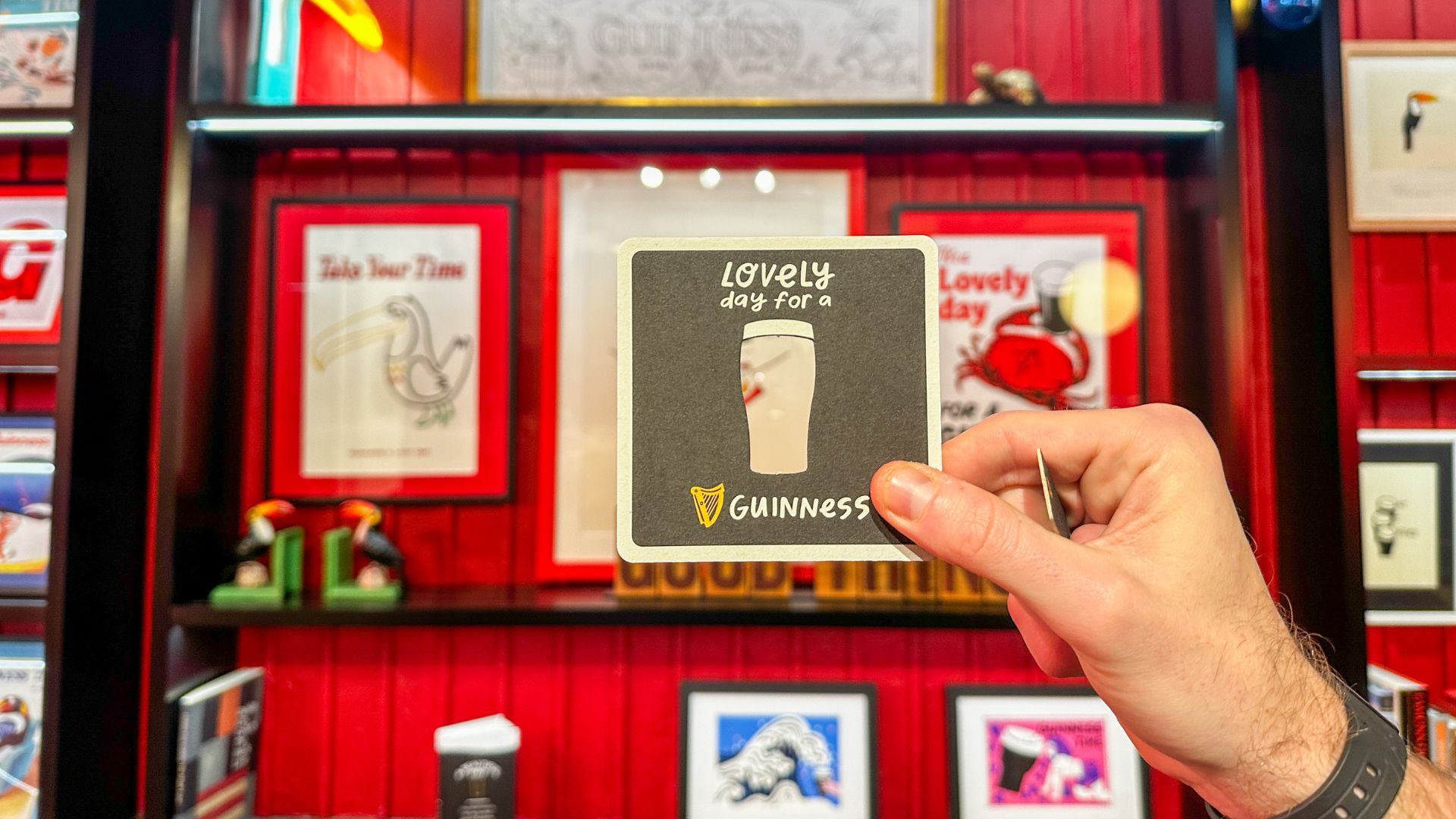 A hand holding a Guinness beer mat reading “Lovely day for a Guinness,” with branded artwork displayed on a red wall behind.