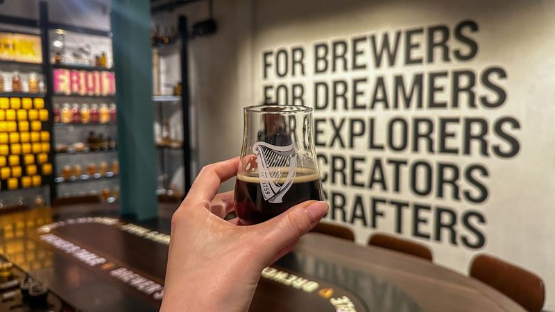 A Guinness tasting glass held inside a modern tasting room at the Guinness Open Gate Brewery, with branded wall text in the background.