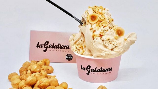 A pink ice cream cup filled with a giant scoop of soft serve ice cream with nuts on top and a black spoon.
