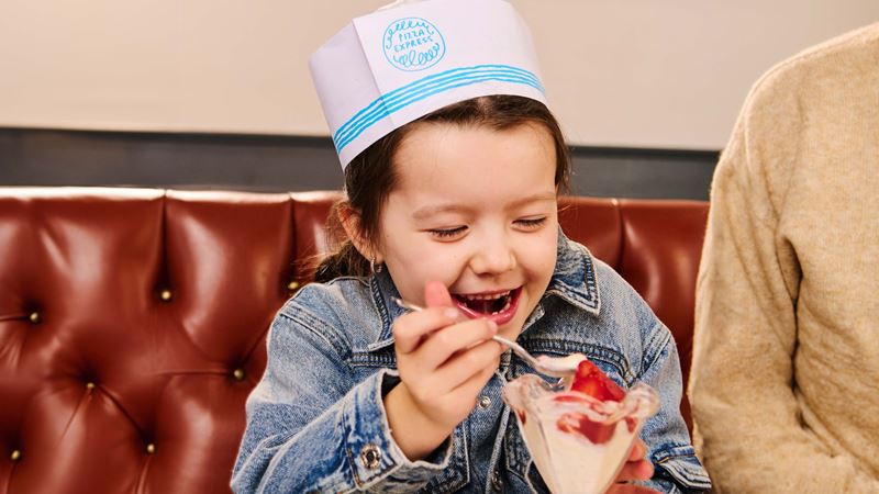 A smiling child wearing a PizzaExpress chef hat eating a strawberry ice cream sundae while sitting in a restaurant booth. 
