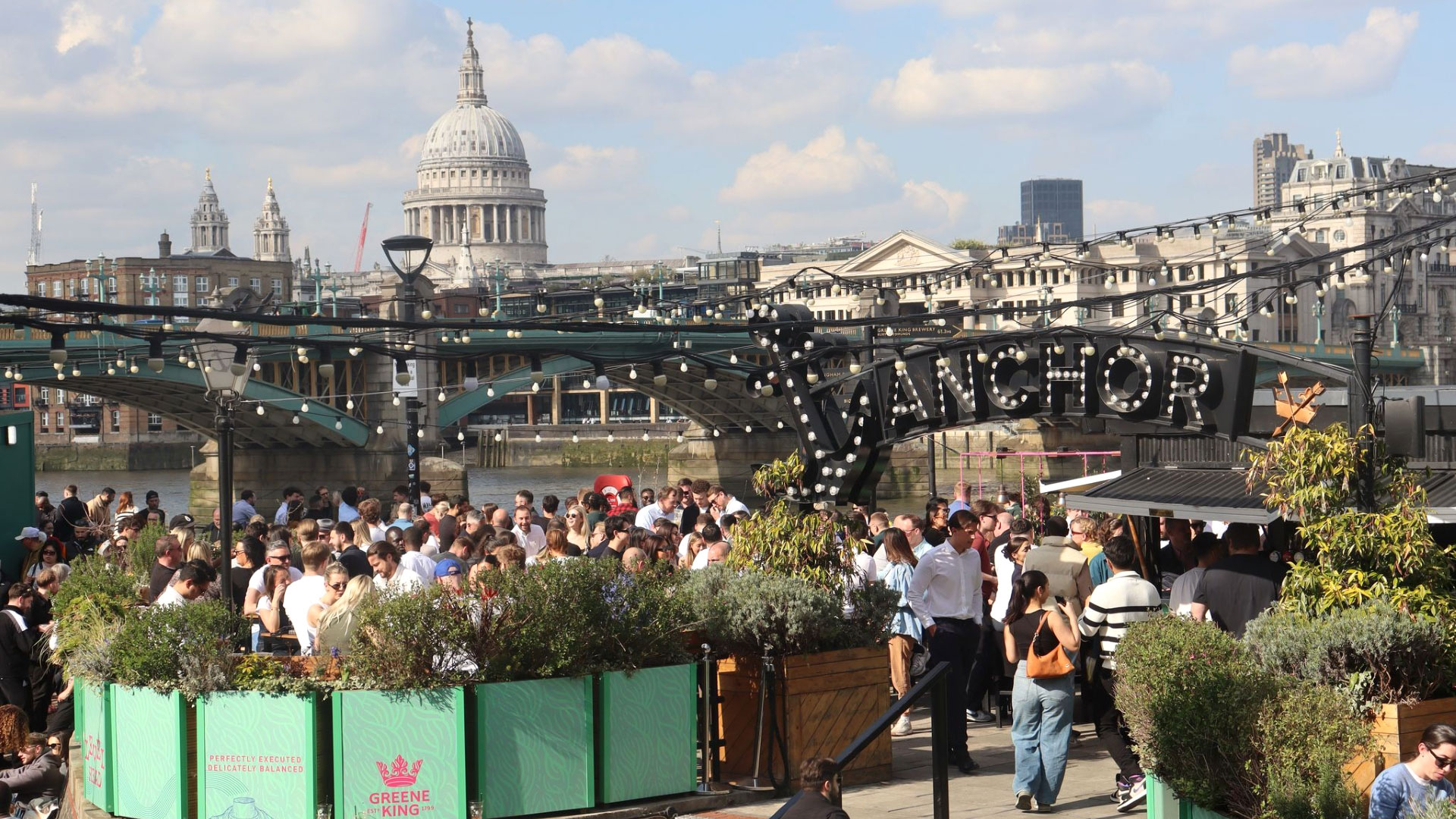 A photo of the riverside garden at Anchor Bankside with crowds of people sat down surrounded by foliage on a bright sunny day with a view of London Bridge and St Paul's Cathedral in the background