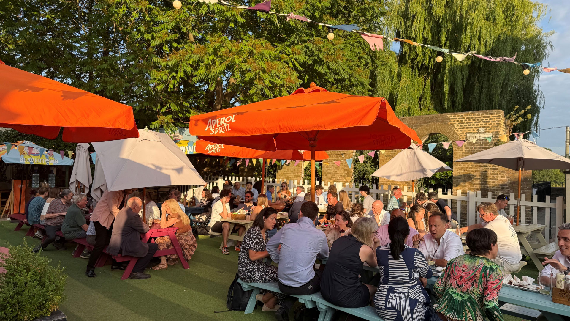 Benches full of people enjoying drinks on a sunny afternoon with bunting and parasols overhead and trees in the distance
