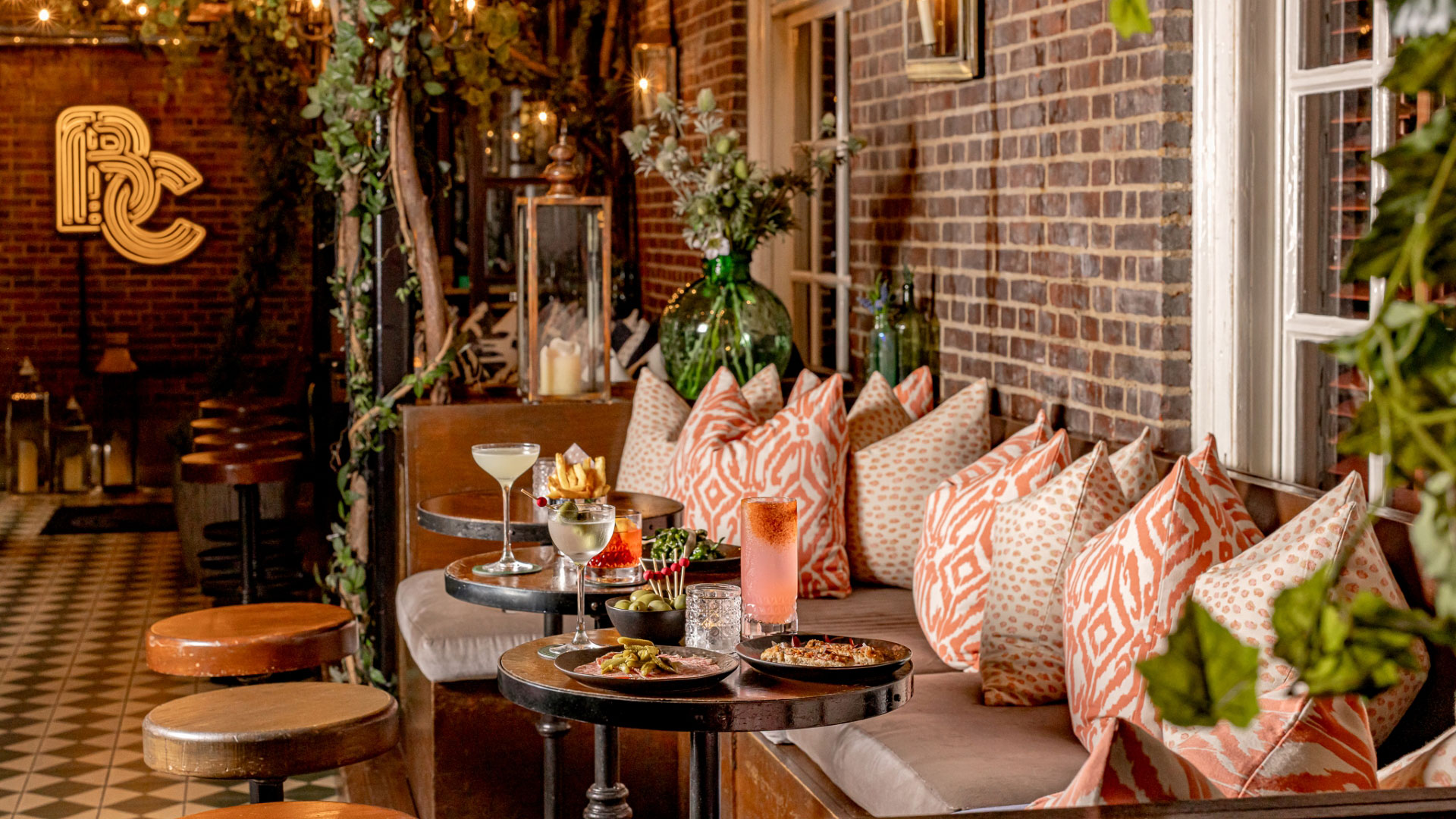 A photo of The Bloomsbury Club bar with comfy sofa seating filled with pillows with stools opposite and tables covered in cocktails and small plates 