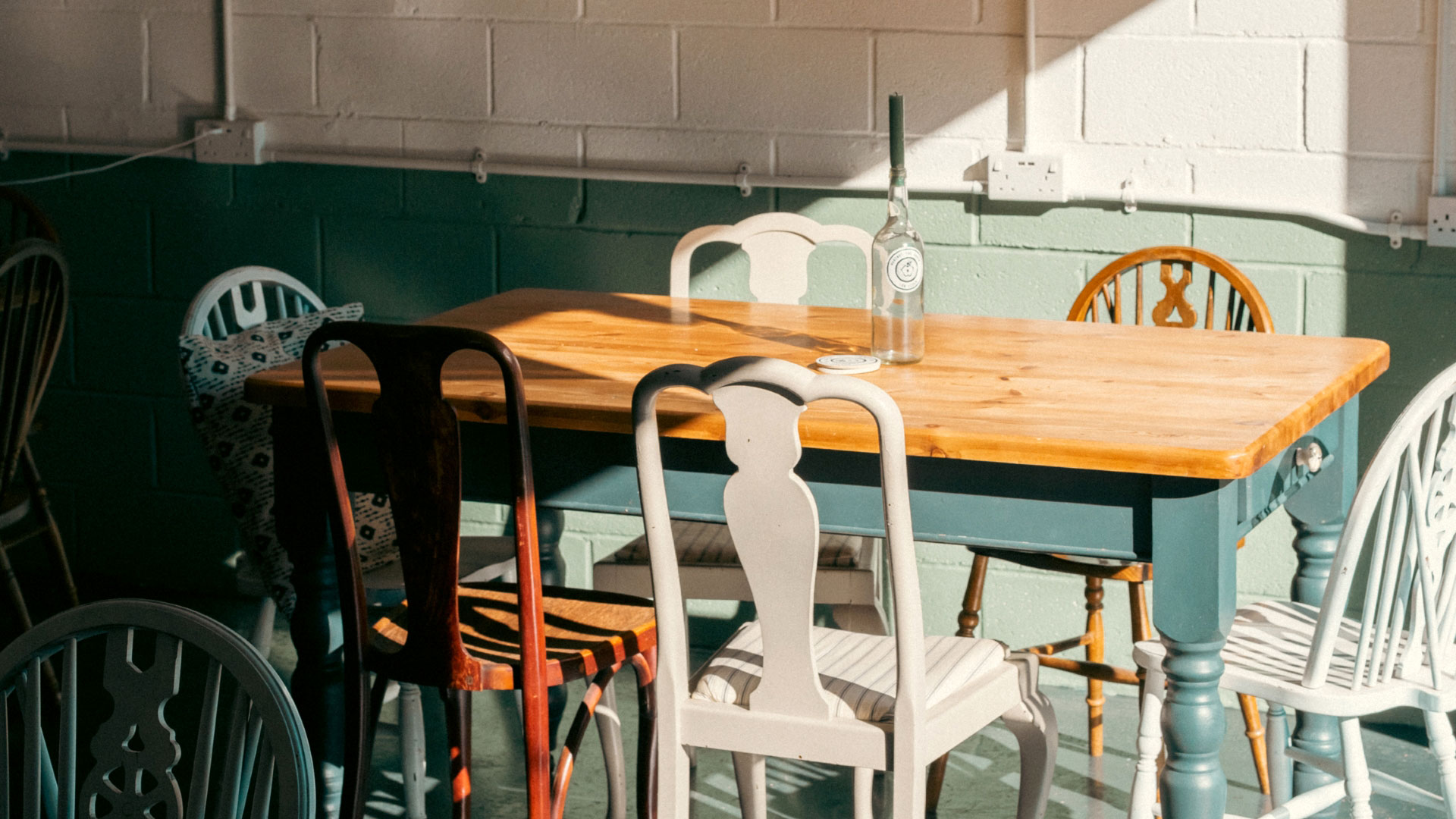 A photo of a wooden table at Against The Grain Cidery taproom with mismatching vintage chairs in different colours and a candlestick inside an empty wine bottle on the table with a ray of sun catching the walls
