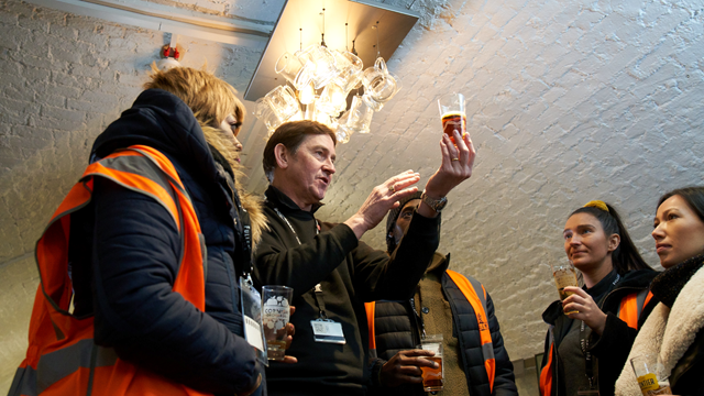 A tour guide holds up a glass of freshly brewed beer while talking to a group wearing orange hi-vis jackets on a Fullers Brewery tour