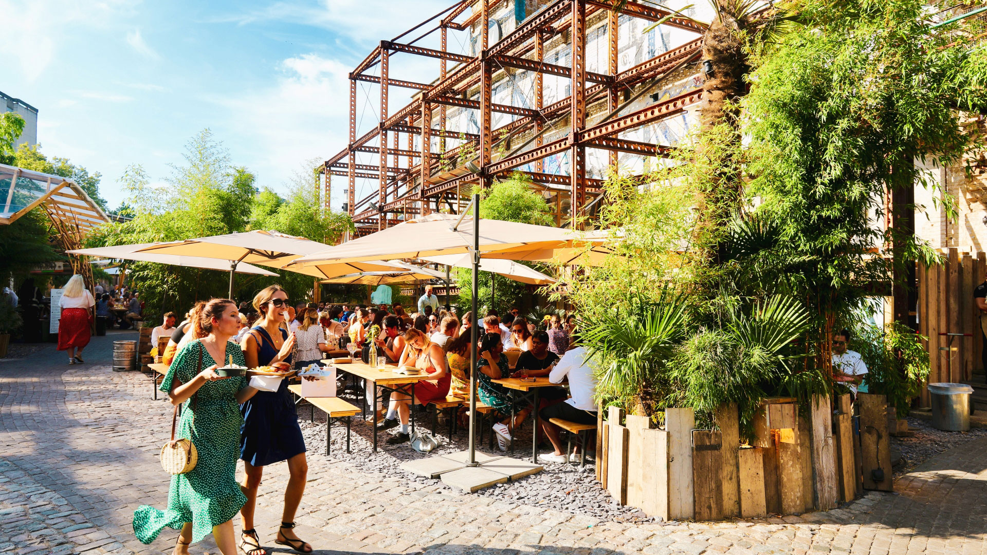 A photo of two female friends wearing dresses walking along a path on a bright sunny day at Mercato Metropolitan with people sat on picnic benches and parasols and plants in frame