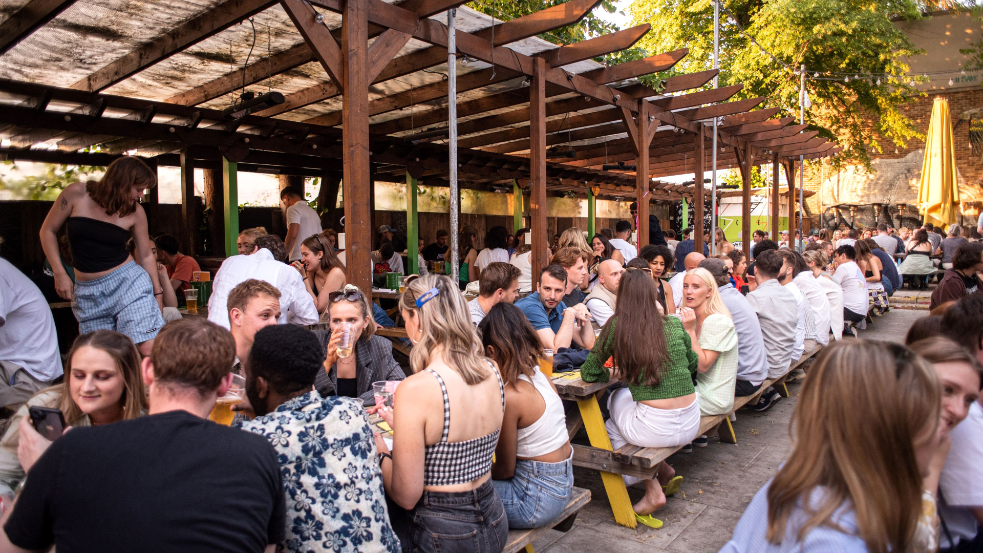 A photo of crowds of people sat on picnic benches in the Duke of Edinburgh's outdoor beer garden with wooden canopies and greenery in the background as the sun shines down