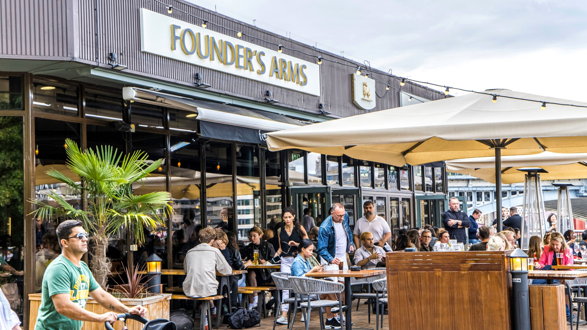 A photo of the Founder's Arms pub on the Thames with benches filled with people and parasols overhead