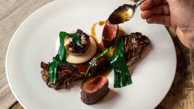 Treat yourself to a Michelin-starred pub feast at The Harwood Arms in Fulham. Credit: Lateef Okunnu. Image courtesy of The Harwood Arms. A photo of a muntjac with ale dish at The Harwood Arms featuring medium rare slices of muntjac, wilted wild garlic leaves and a man's hand dripping an ale sauce over the dish with a spoon