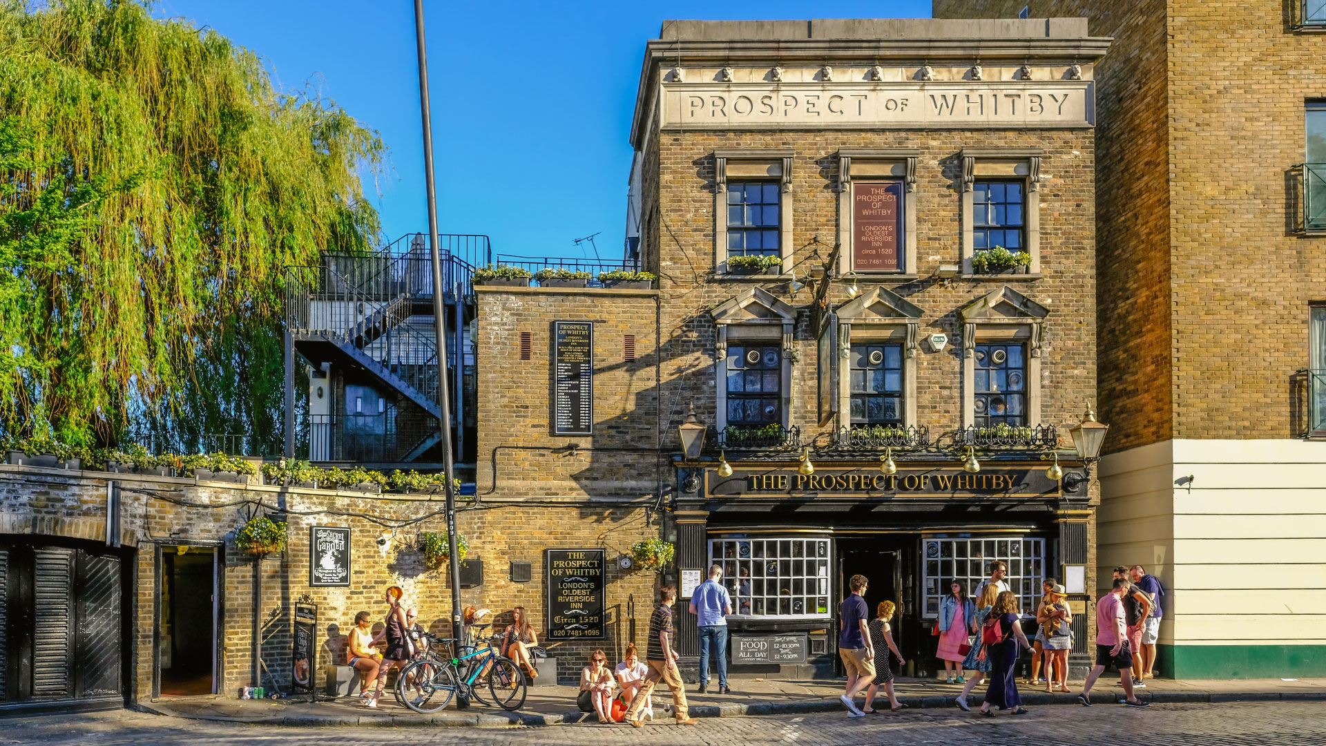 A photo of the exterior of the Prospect of Whitby pub set in a large Victorian building with hanging baskets filled with flowers and people stood outside the front