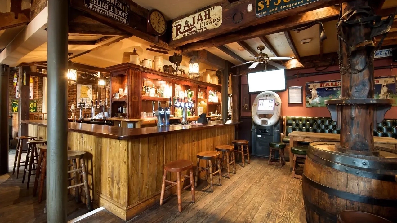 The wooden bar area at The Alexandra pub in Clapham, with stools, drinks and a TV screen in the background.