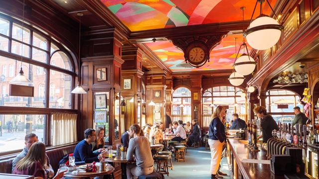 The interior of The Audley pub in Mayfair, with wood panelling on the walls, a colourful painted ceiling and traditional bar, with pub-goers sitting on stools and benches.
