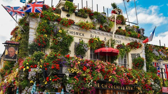 The exterior of The Churchill Arms, which is covered in hanging baskets, shrubs and flowers, with a pub sign showing Winston Churchill's face.