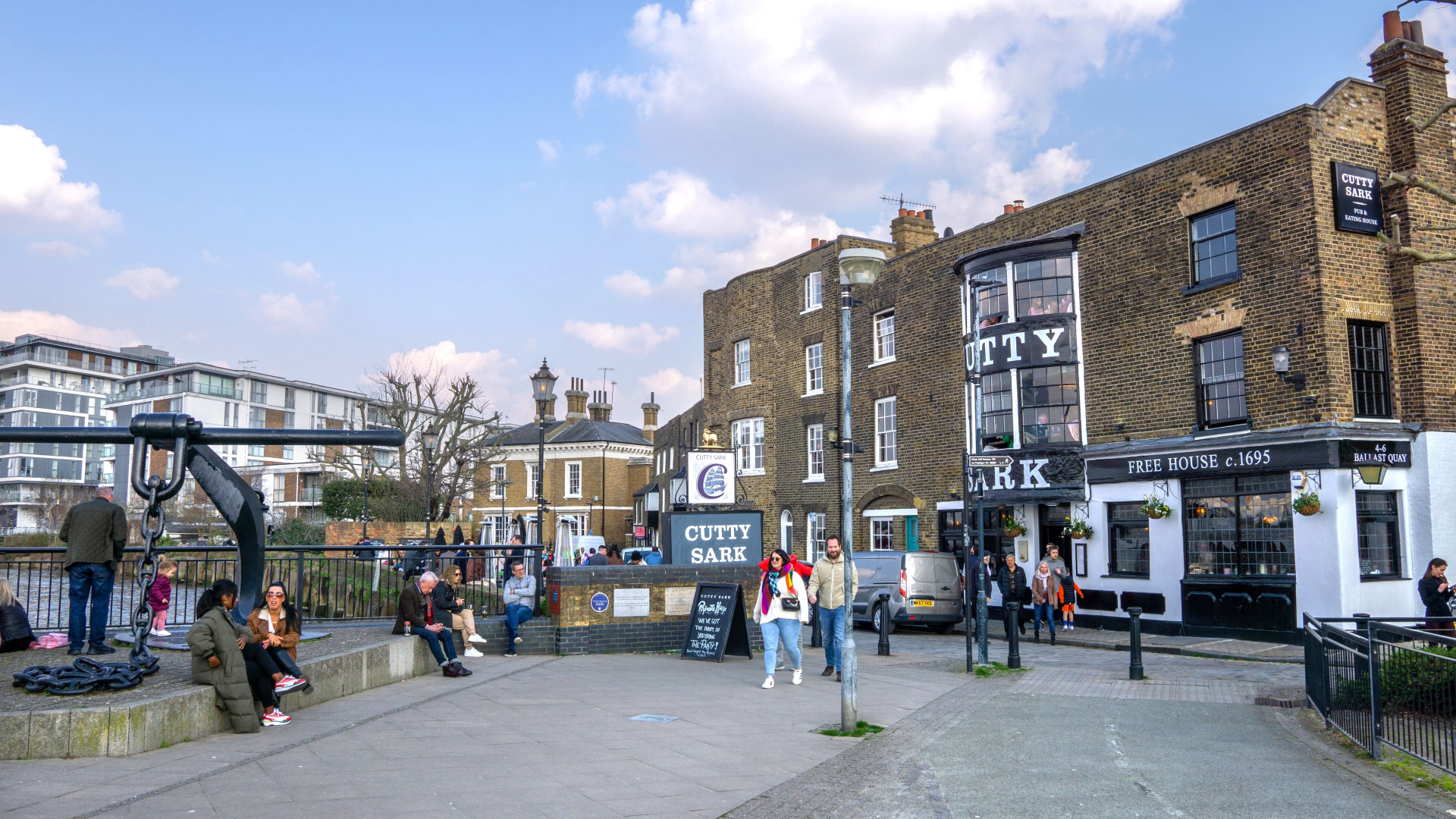 A photo of the exterior of the Cutty Sark pub in Greenwich with people walking on the street in front, a giant anchor sculpture and a snippet of the Thames on the left of the image