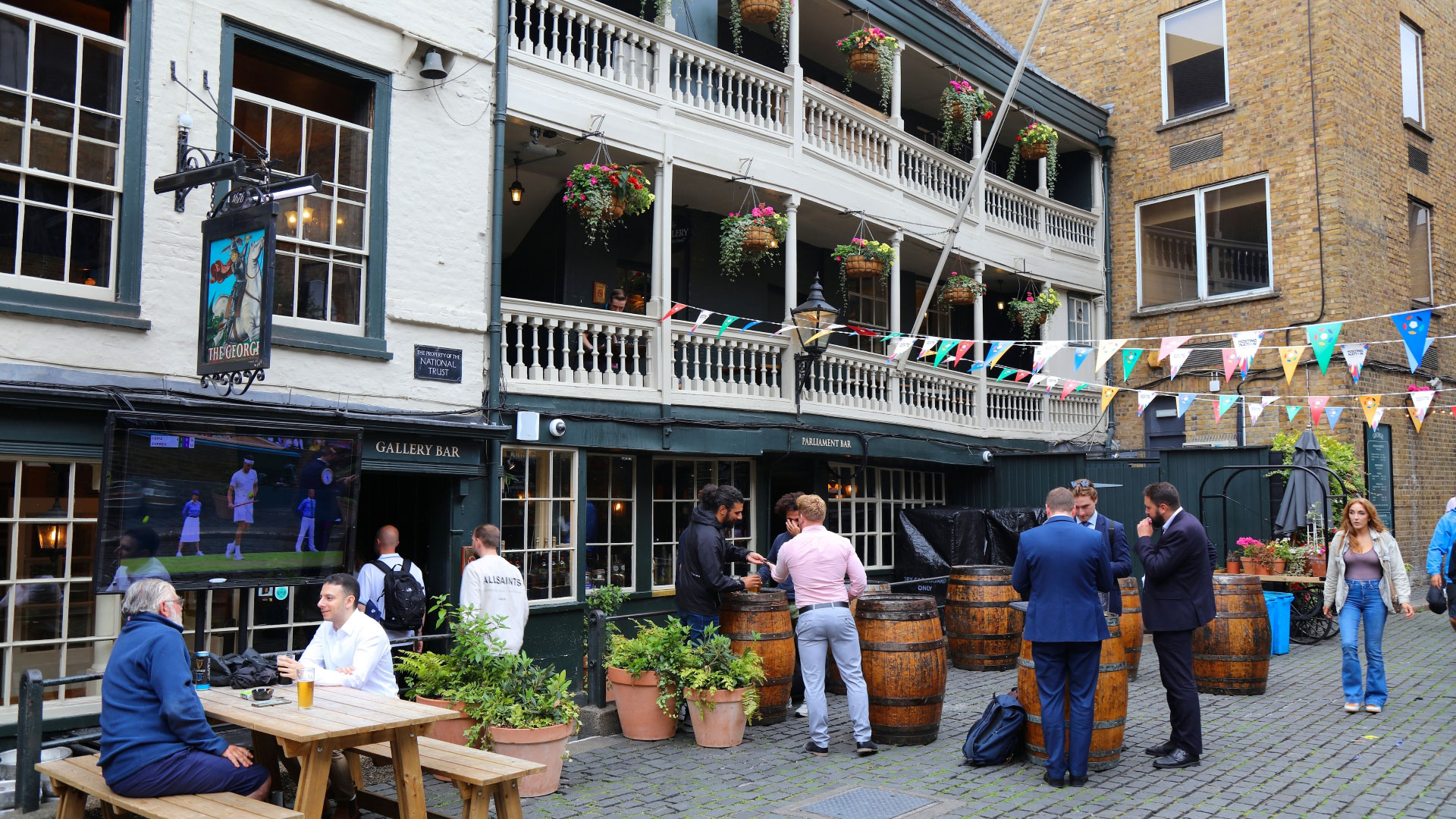 People sit on and stand next to wooden tables in front of The George Inn with its galleried exterior and hanging flower baskets