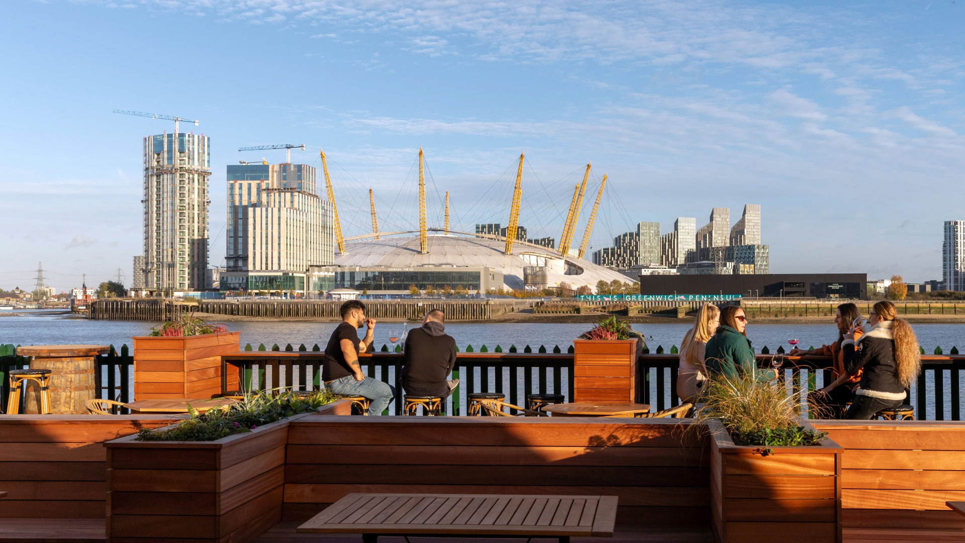 A photo of groups of people sat on stools in the riverside garden at The Gun with views of the river Thames and the south bank with The O2 in the distance