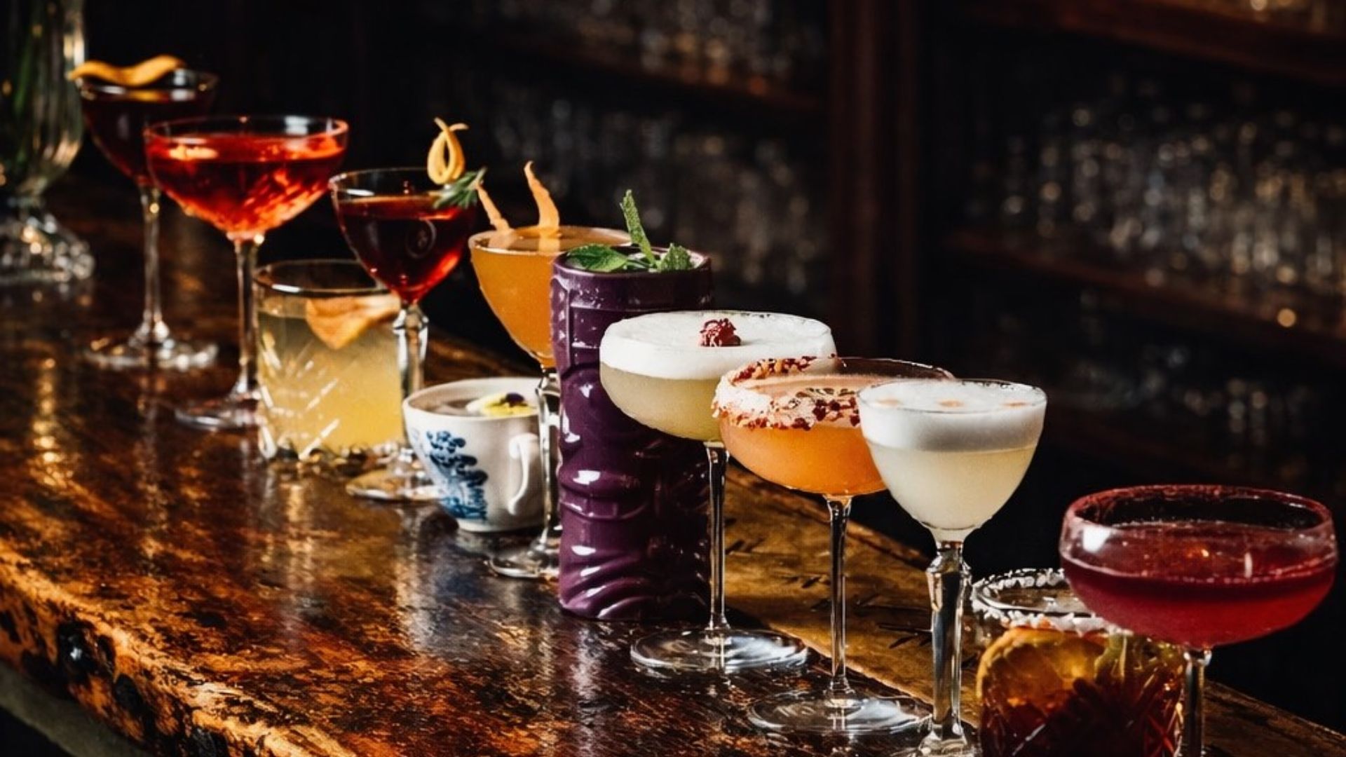 A selection of colourful cocktails lined up on a bar in a dimly lit setting.