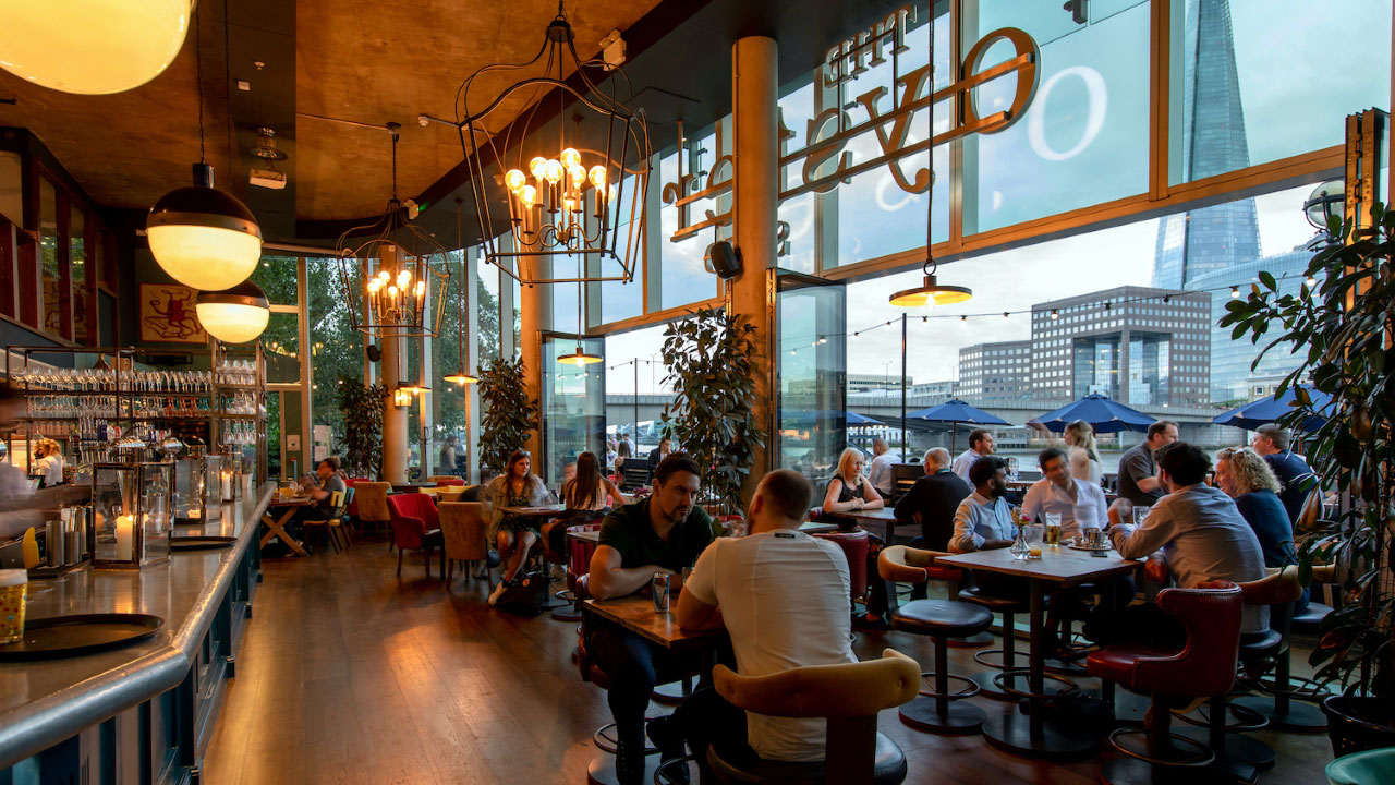 A photo of The Oyster Shed interior with people sat next to floor-to-ceiling windows with views of the south bank of the Thames including The Shard 