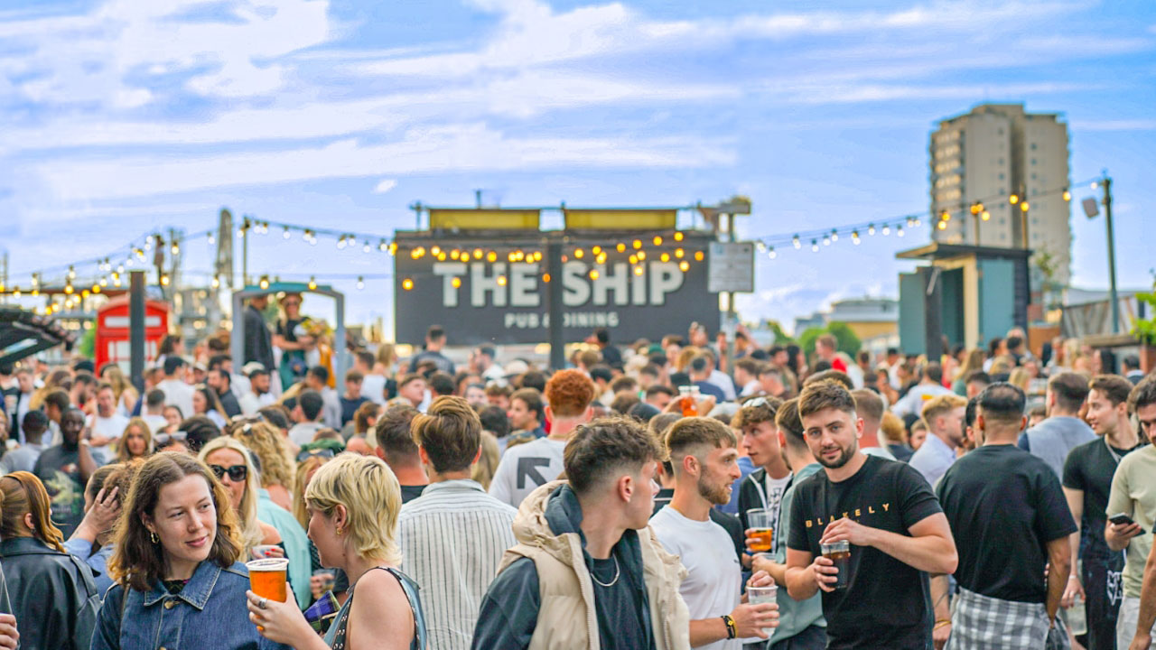 A photo of crowds of people chatting, smiling and with drinks in hand in the garden at The Ship with fairy lights and a blue sky above