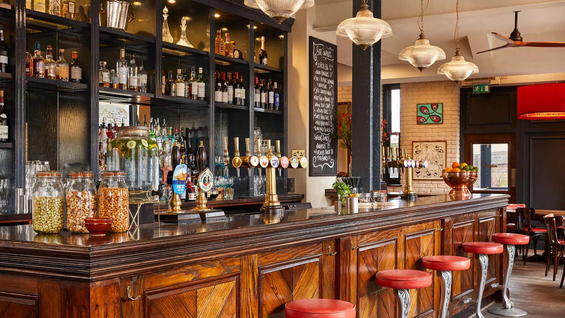 Seek out London's cosiest pubs, such as The Stonhouse in Clapham. Credit: The Stonhouse. Image courtesy of Three Cheers Pub Co. Inside a cosy pub in London, with a wooden bar area and drinks lined on shelves in the background.