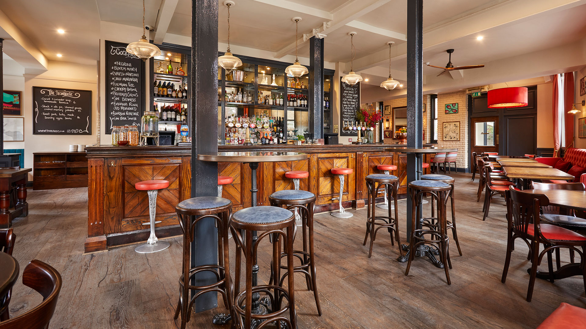Inside The Stonhouse pub in Clapham, featuring wooden floors and tables, red and blue seats, a wooden bar area with drinks lined on shelves in the background.