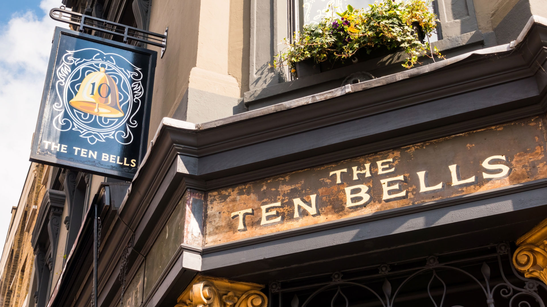 A photo of The Ten Bells signage outside the front of the pub with a blue placard hanging off the side of the building featuring a gold bell