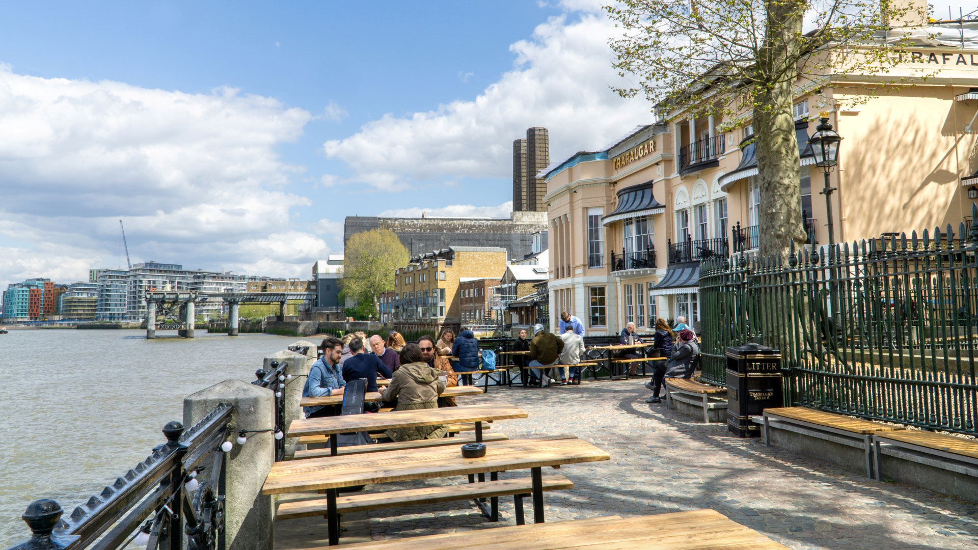 A photo of the exterior of the Trafalgar Tavern with people sat on the riverside benches with the Thames in front of the pub
