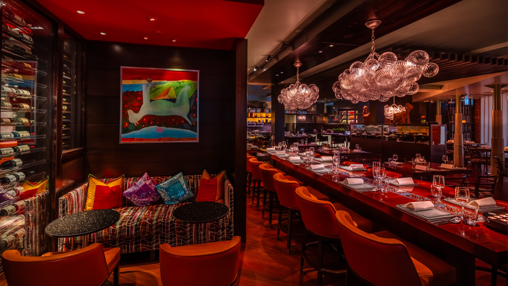 Darkly lit Amaya restaurant interior, featuring red carpeting, brown walls, a lounge area with colourful sofa, red seats and cushions, and a counter with red bar stools, plates and glasses.