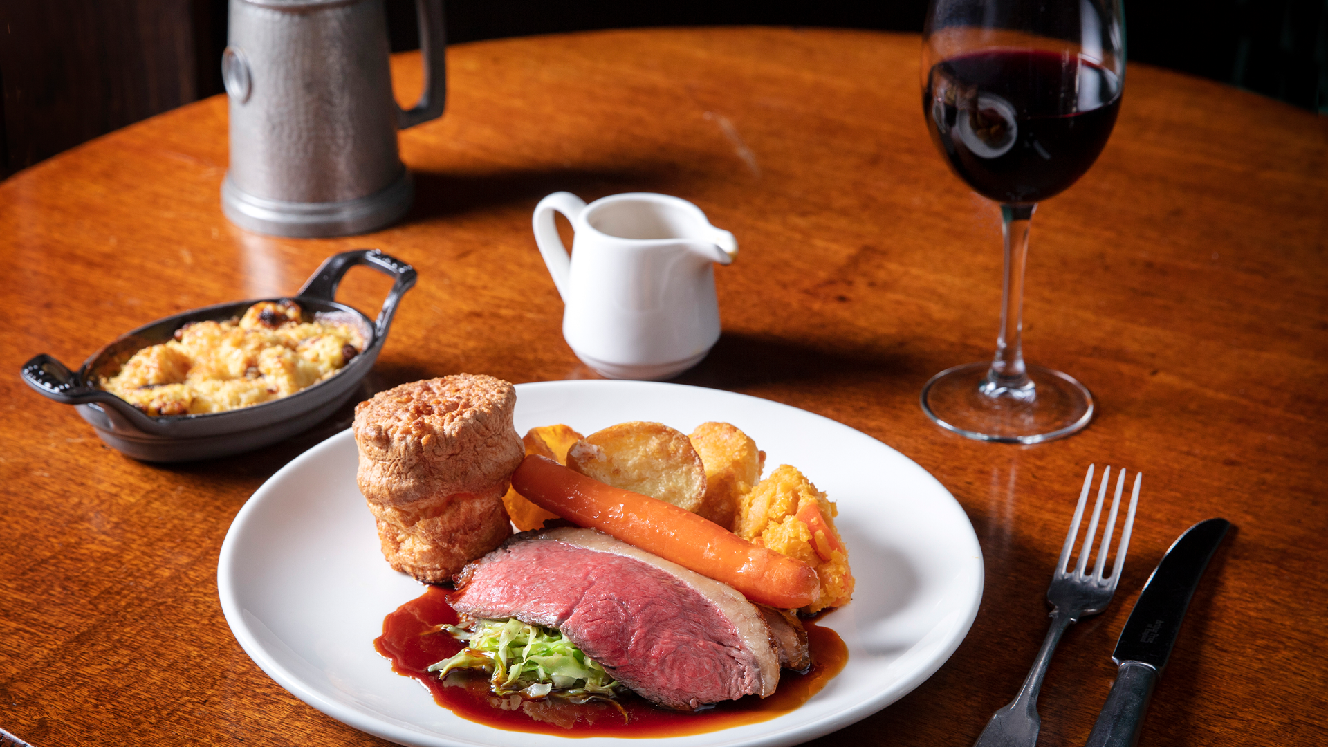 A plate of roast beef, vegetables, Yorkshire pudding and gravy, a glass of wine and cutlery.