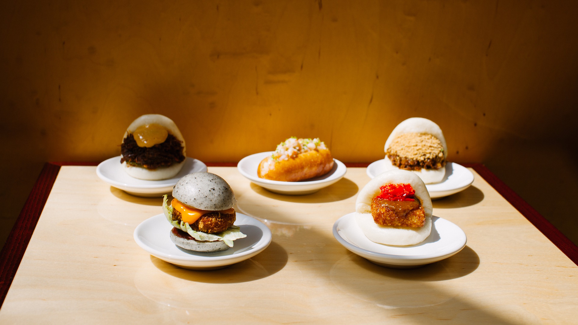 Four small plates, each with fluffy bao buns with different fillings, on a light-coloured wooden table top.