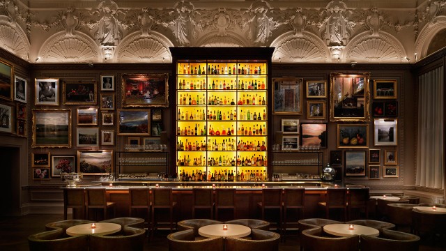 A wall covered in framed artwork inside a dimly lit room with a bright yellow shining bar stocked to the brim