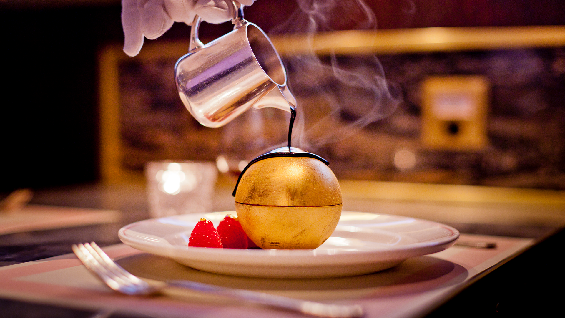 Hot chocolate being poured over a dessert hidden inside a golden chocolate dome, with raspberries on the side on a white plate at Bob Bob Ricard in London