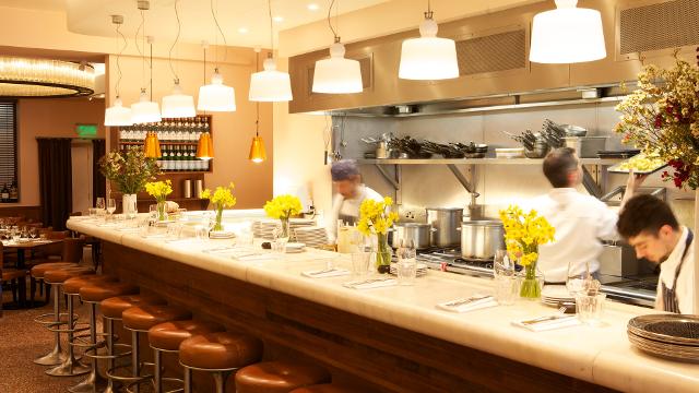 A white bar with brown stools and chefs cooking.