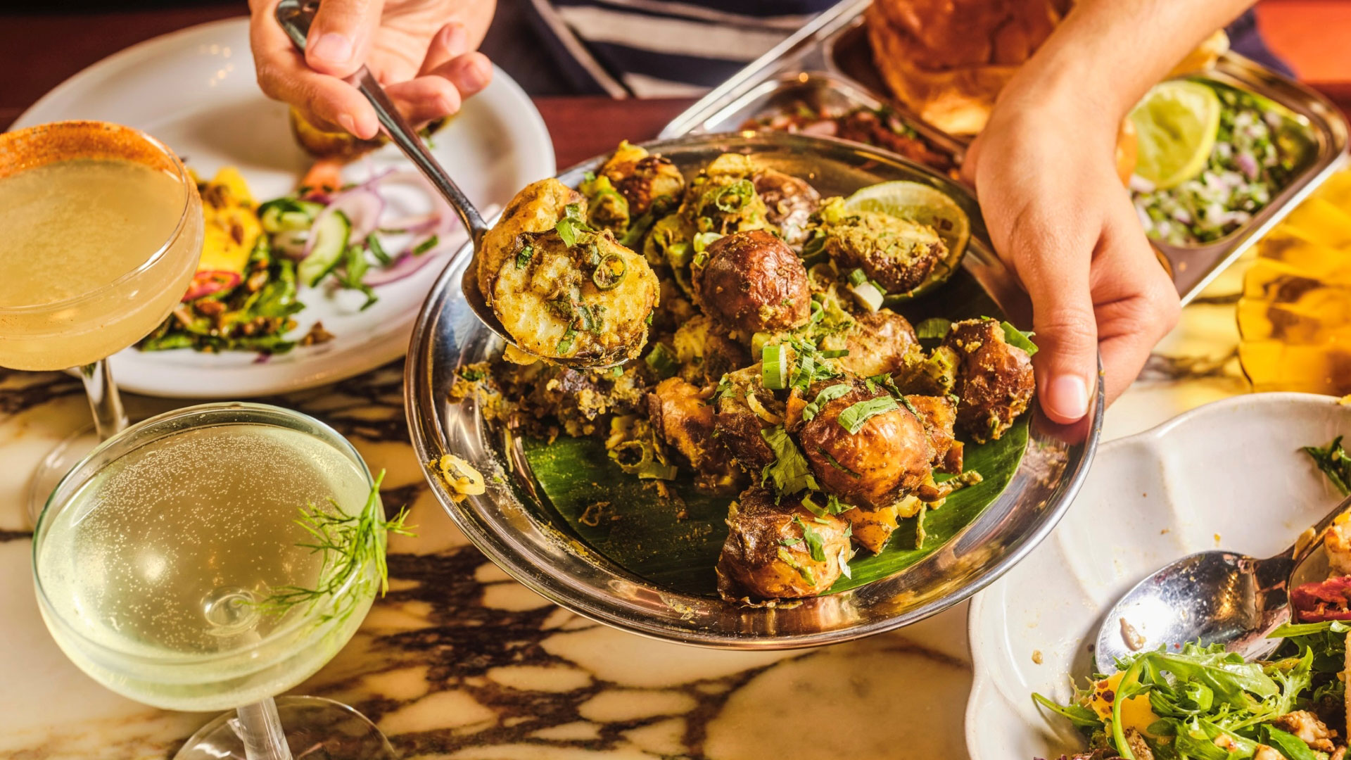 A bird's eye photo of someone holding a dish of gunpowder potatoes at Dishoom with other dishes and cocktails on a table underneath