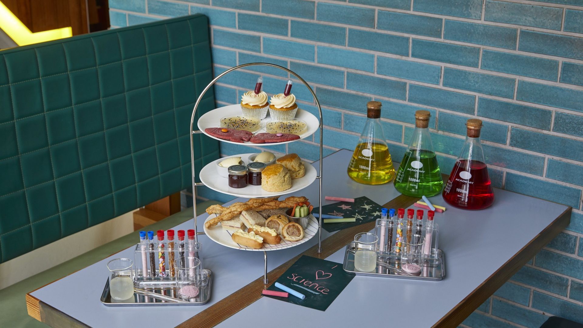 A science-themed afternoon tea with cakes, sandwiches and colourful drinks presented alongside laboratory-style glassware and test tubes.