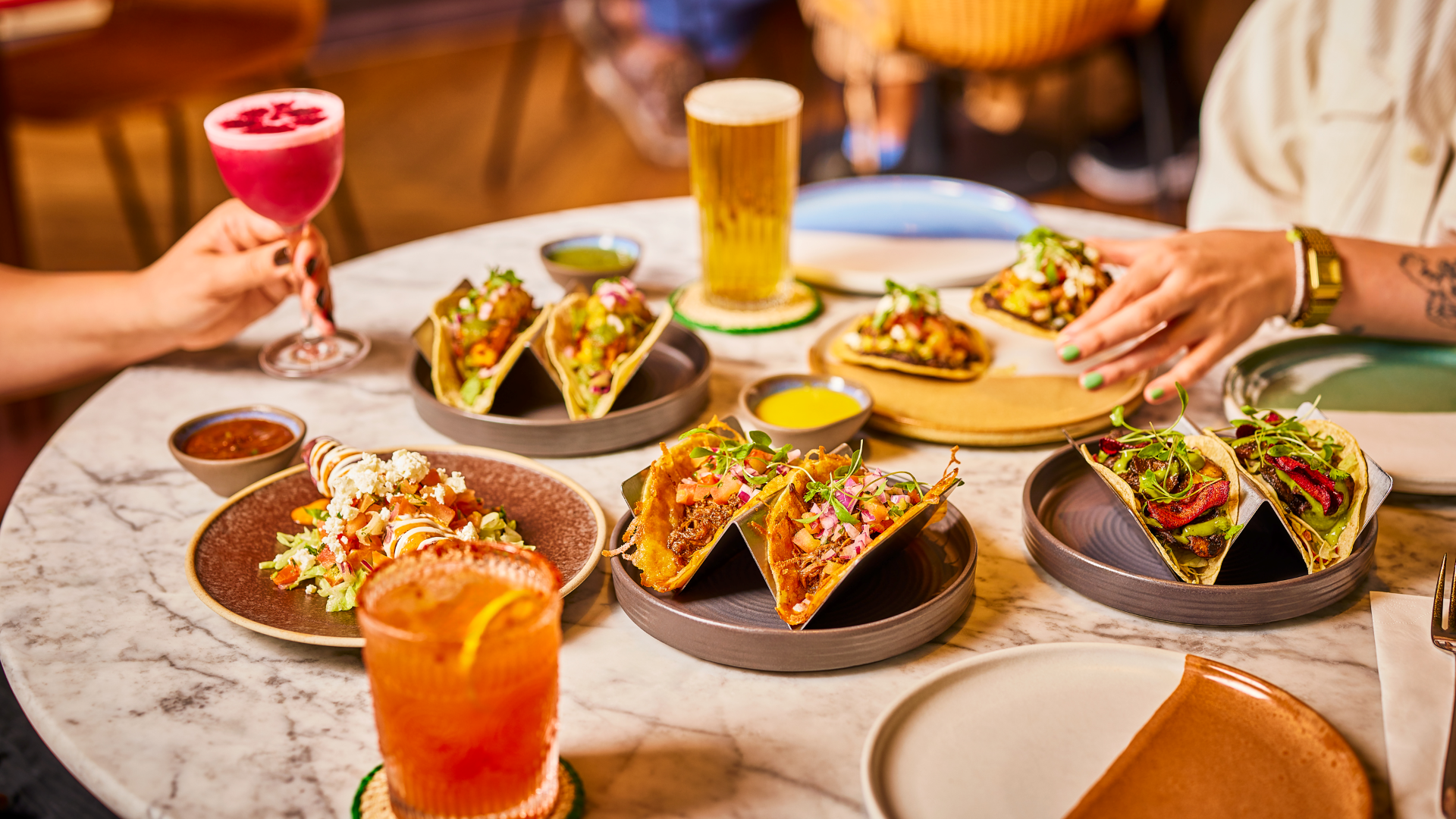 A white marble table with plates of colourful Mexican food including tacos and enchiladas, cocktails and beer