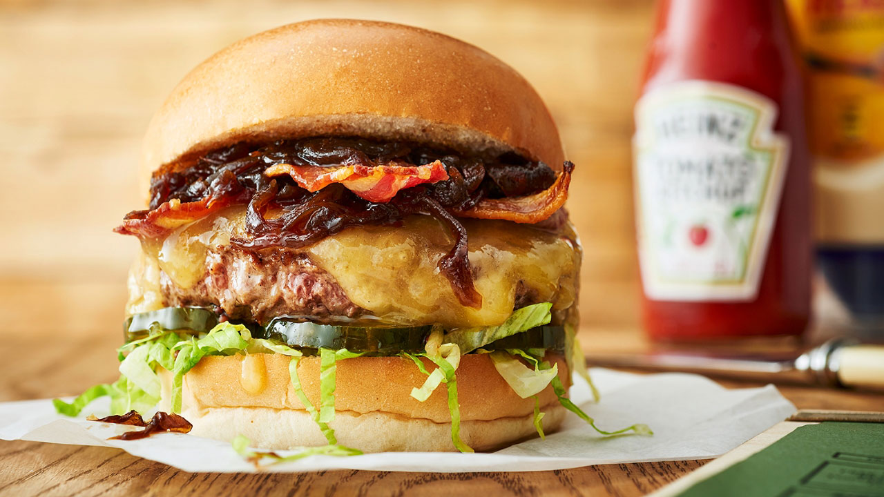 A close up photo of a burger at Honest Burgers with a juicy beef patty, melted cheese, salad and crispy bacon with a bottle of tomato ketchup in the background