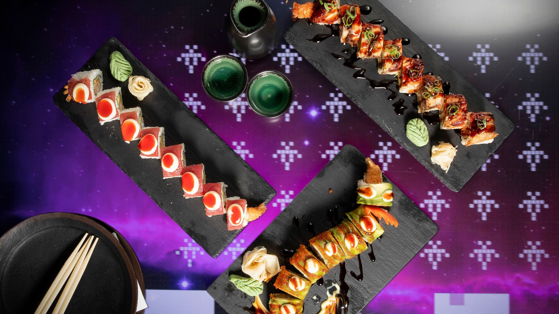 Assorted sushi rolls arranged on black serving plates with dipping sauces and drinks on a table with a digital interactive surface.