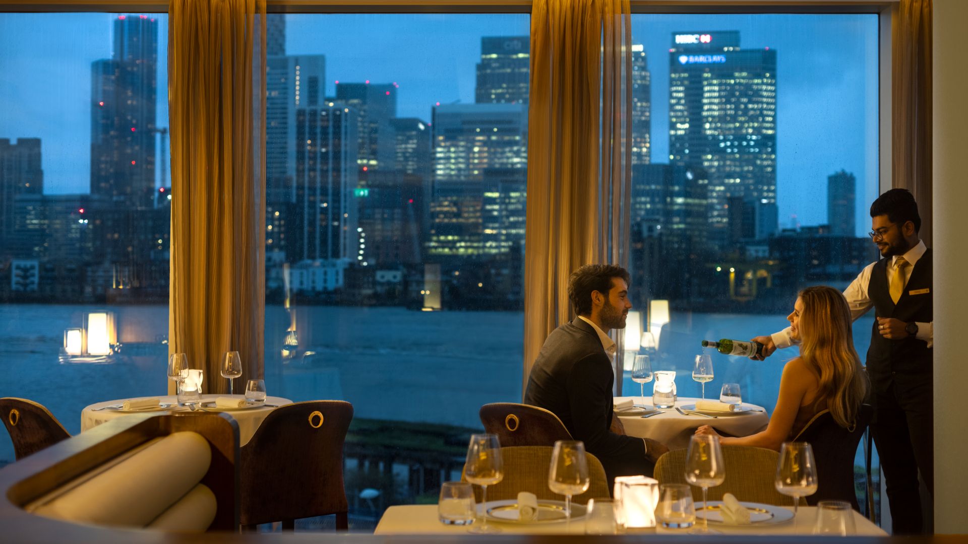 A cosy restaurant with ambient lighting and a couple sat in front of the wall of windows with a view over the London city skyline at dusk.