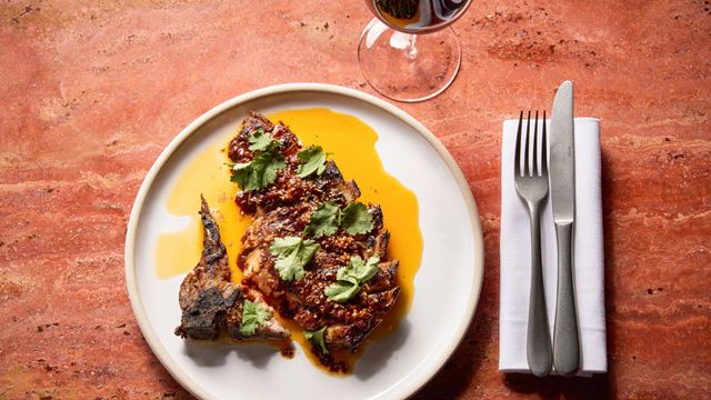 Aerial shot of pork chop in jus on a warm orange marble table with silver cutlery and a glass of red wine.