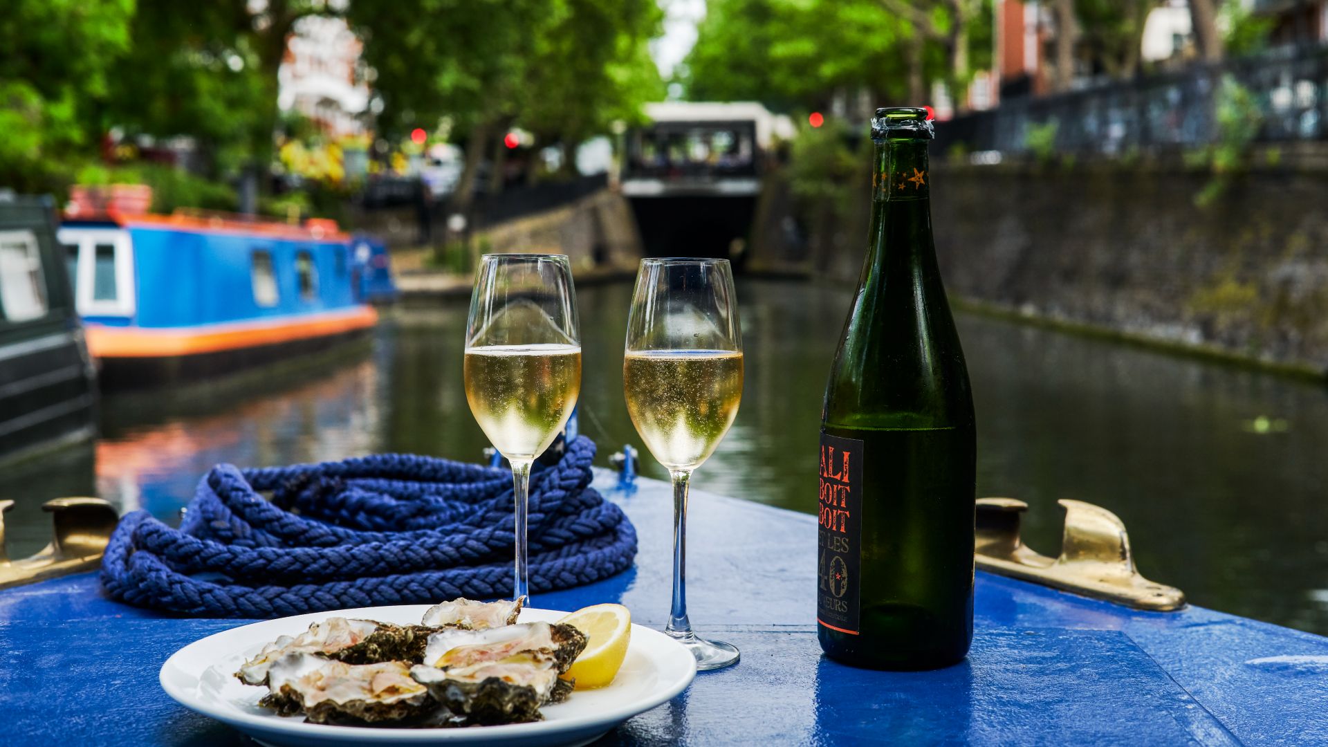 A landscape view of a seafood platter and bottle of wine on a barge, with the canal in the background blurred.