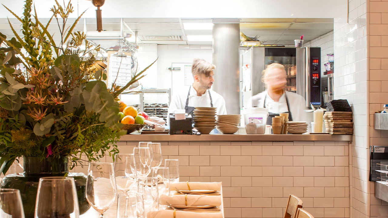 A photo of two chefs in the kitchen from the dining area with stacks of bowls and plates on the counter and a table covered in crockery, glassware, cutlery and flowers in the foreground
