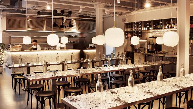 Main space inside Pastaia restaurant in Soho, with long tables, wooden stools, low-hanging lighting and bottles of water on the tables.
