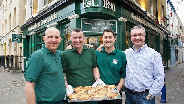 Four men dressed in green shirts stand in front of a london pie shop holding a tray of pies.