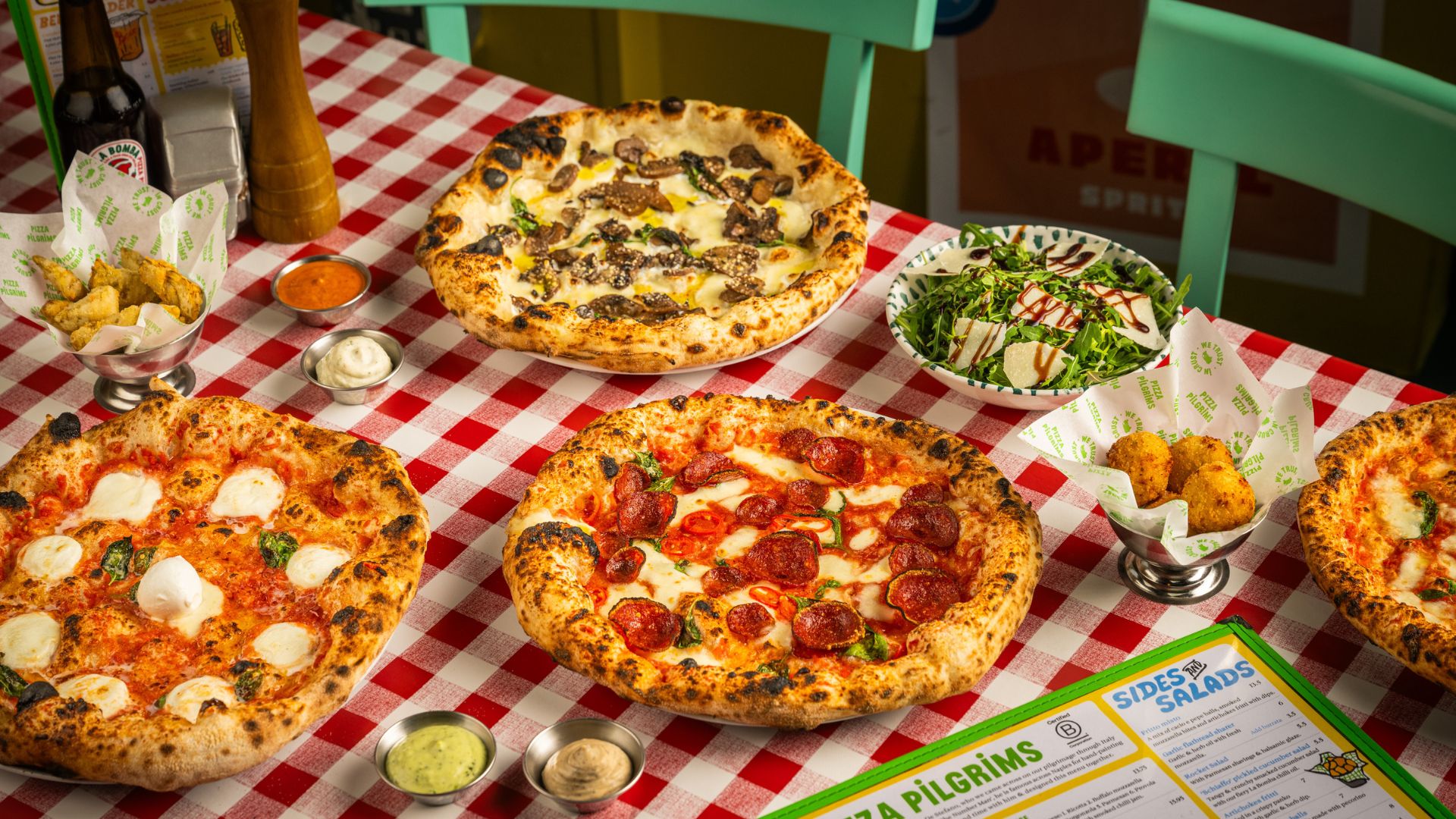 Gather the family around the table at Pizza Pilgrims, where fluffy Neapolitan pizzas, crispy sides and colourful salads make sharing a joyful part of the meal. Image courtesy of MeMo. A spread of Neapolitan pizzas, salads and fried sides served on a red-and-white checkered tablecloth at Pizza Pilgrims.