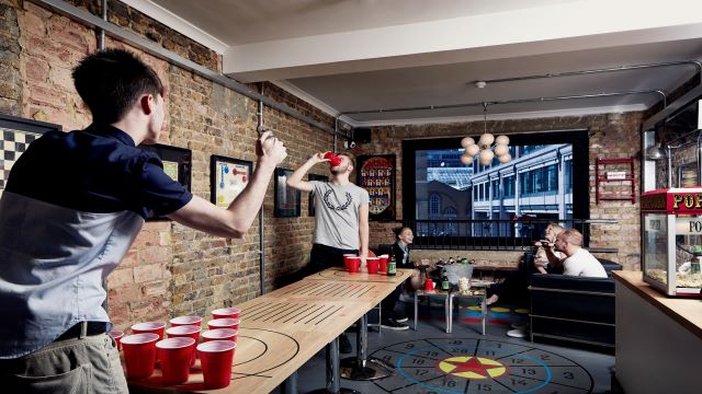 a brick-wall exposed bar lounge, with two people playing beer pong and a group of people enjoying drinks in the background
