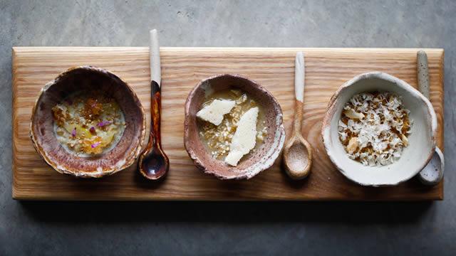 Three bowls filled with different types of porridge and large spoons on a long wooden board.